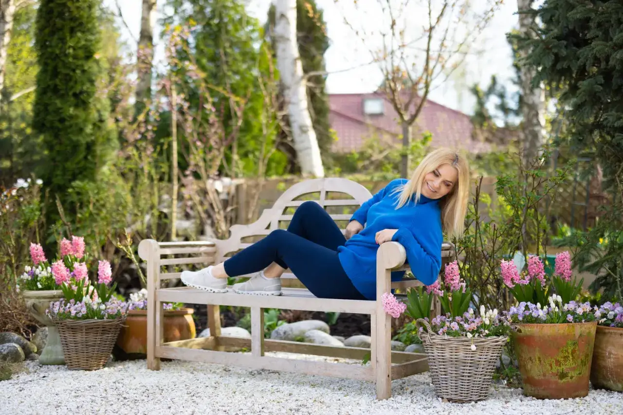 Kobieta w niebieskim swetrze odpoczywa w ogrodzie, otoczona kwitnącymi kwiatami. Zastanawia się, czy bratki są wieloletnie, podziwiając wiosenną zieleń.